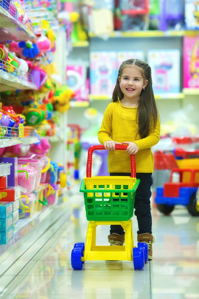 Adorable young girl happily pushing a toy shopping cart in a colorful toy store aisle.